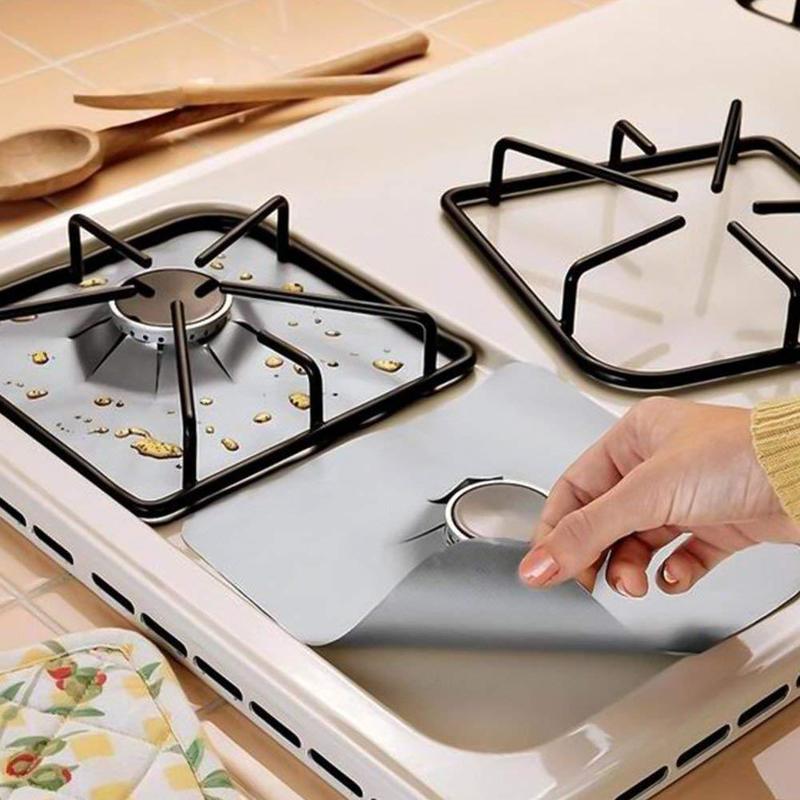 Hand placing gray gas stove burner cover protector on white stovetop with spilled oil and wooden spoons nearby